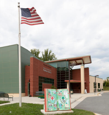 A brick library building with a flagpole, American flag, and an open book sculpture on the front lawn.
