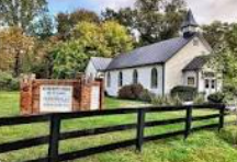 White church with steeple behind a black fence and sign, surrounded by trees and greenery White church with steeple behind a black fence and sign, surrounded by trees and greenery