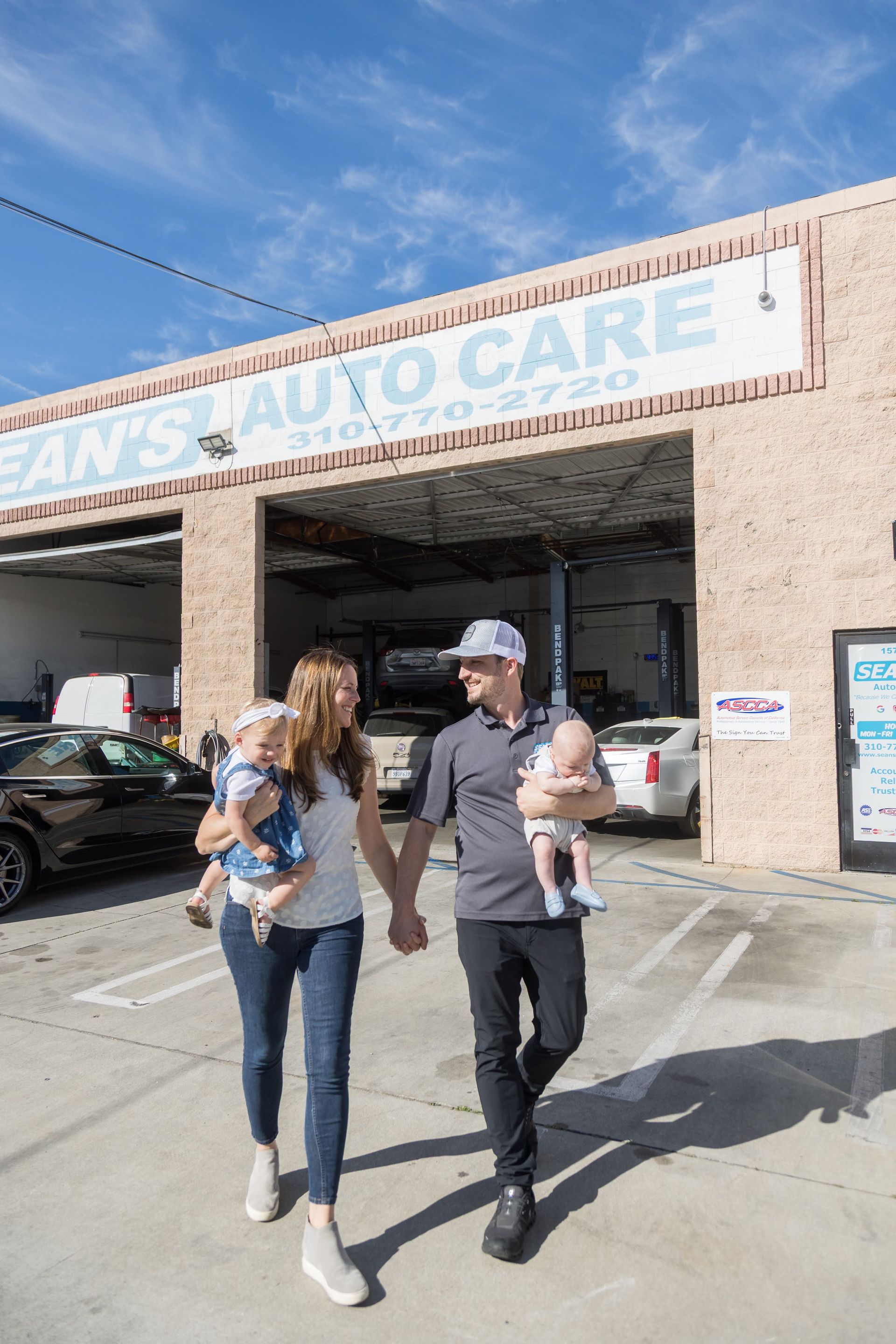 Two people walking outside an auto care shop, carrying a baby and a small dog. | Sean's Auto Care