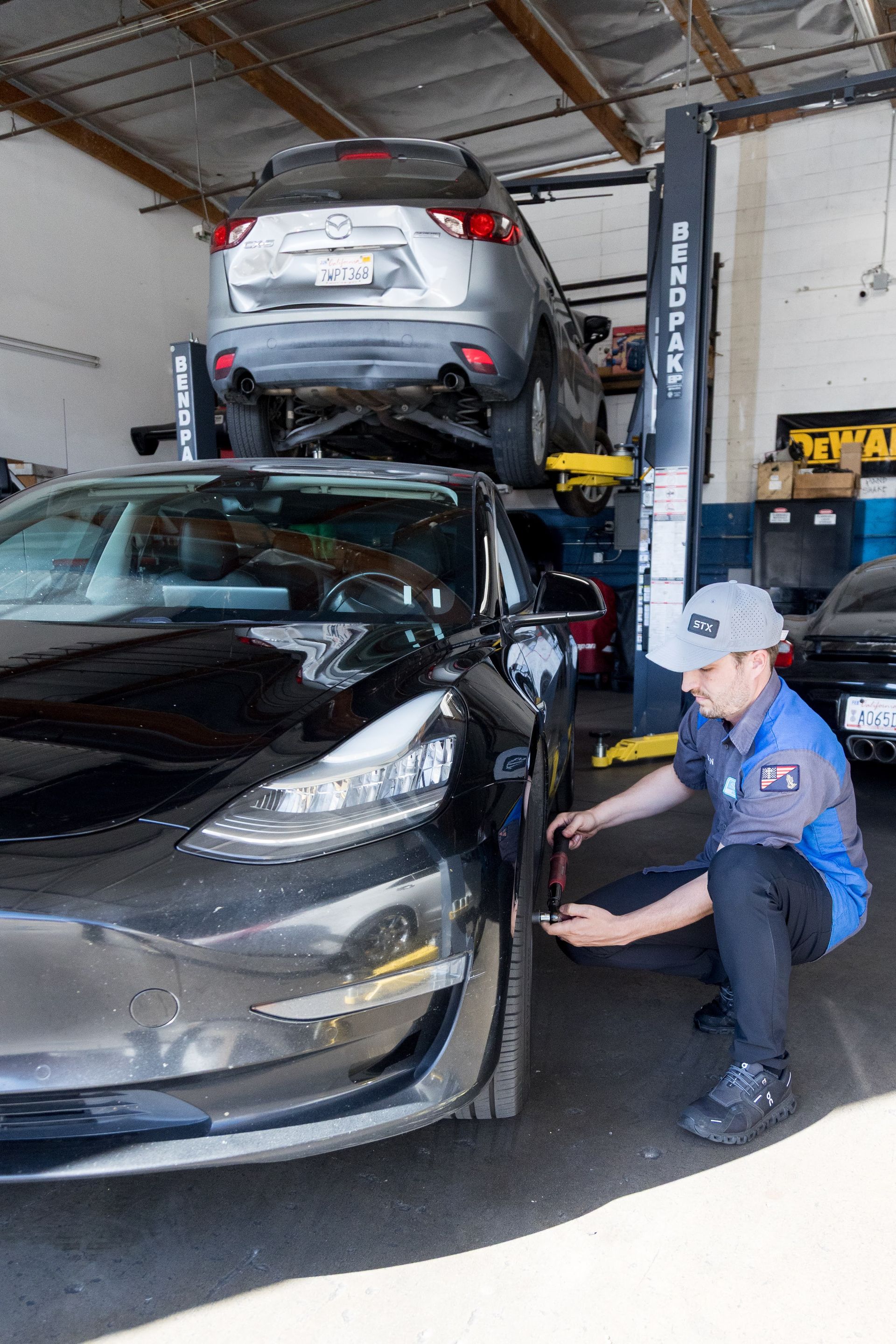 Mechanic in blue uniform inspecting a black car in a garage, with a silver car raised on a lift behind it | Sean's Auto Care