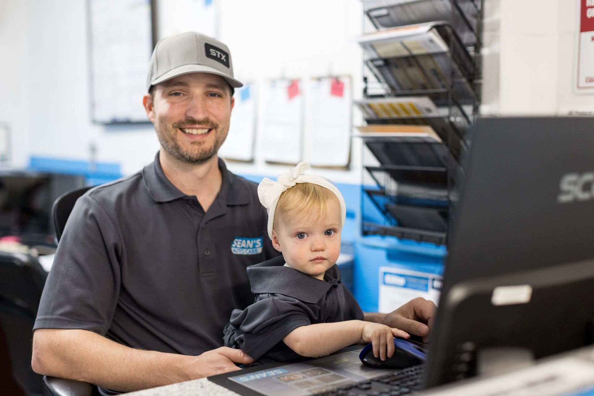 Man and toddler in matching dark shirts using a computer in an office or workspace. | Sean's Auto Care