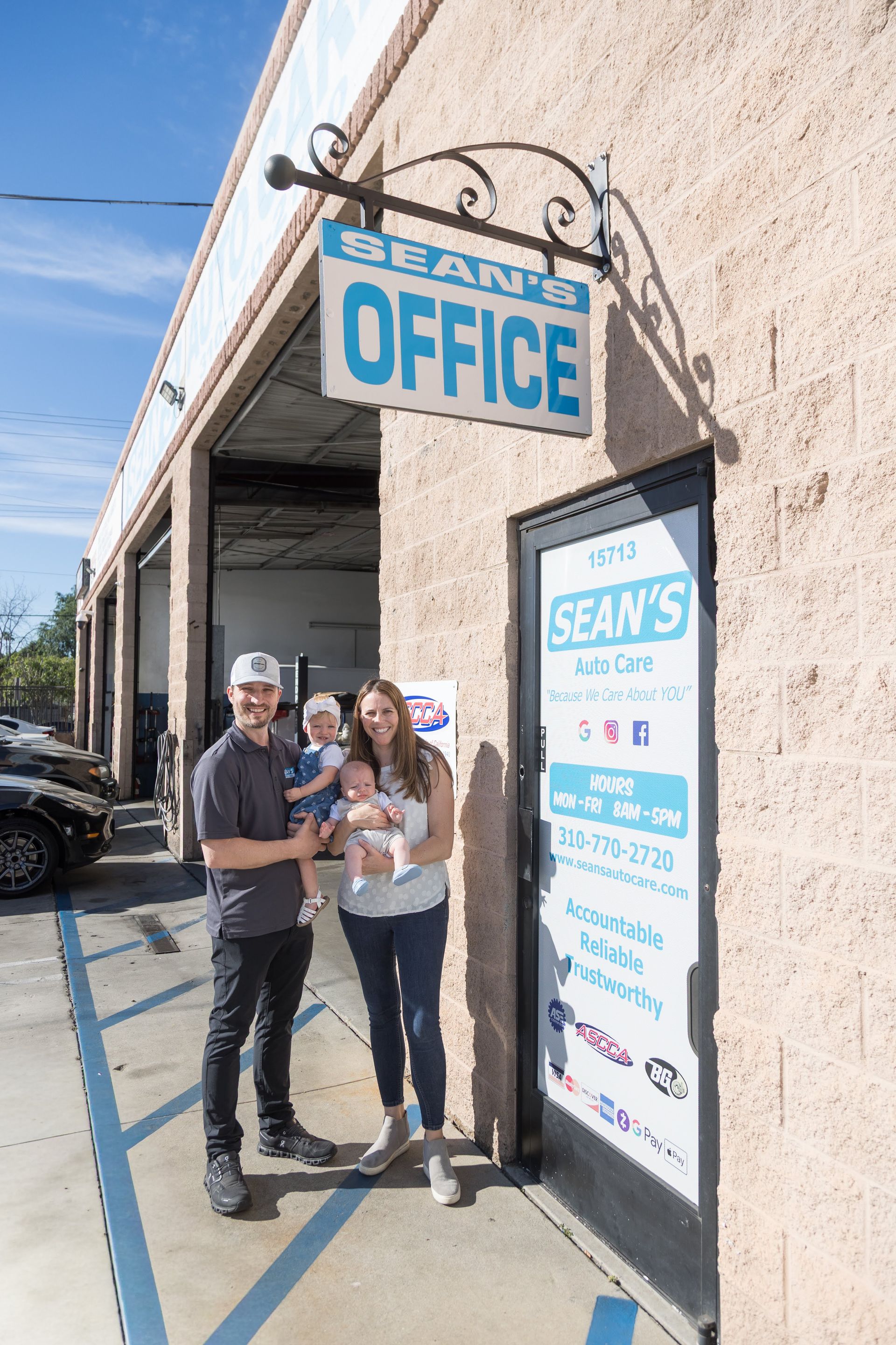 Three people stand outside Sean’s Office storefront, smiling by the entrance sign. | Sean's Auto Care
