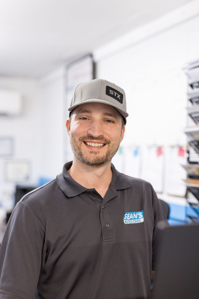 A man in a gray and blue shirt is standing in front of a car in a garage - Sean's Auto Care