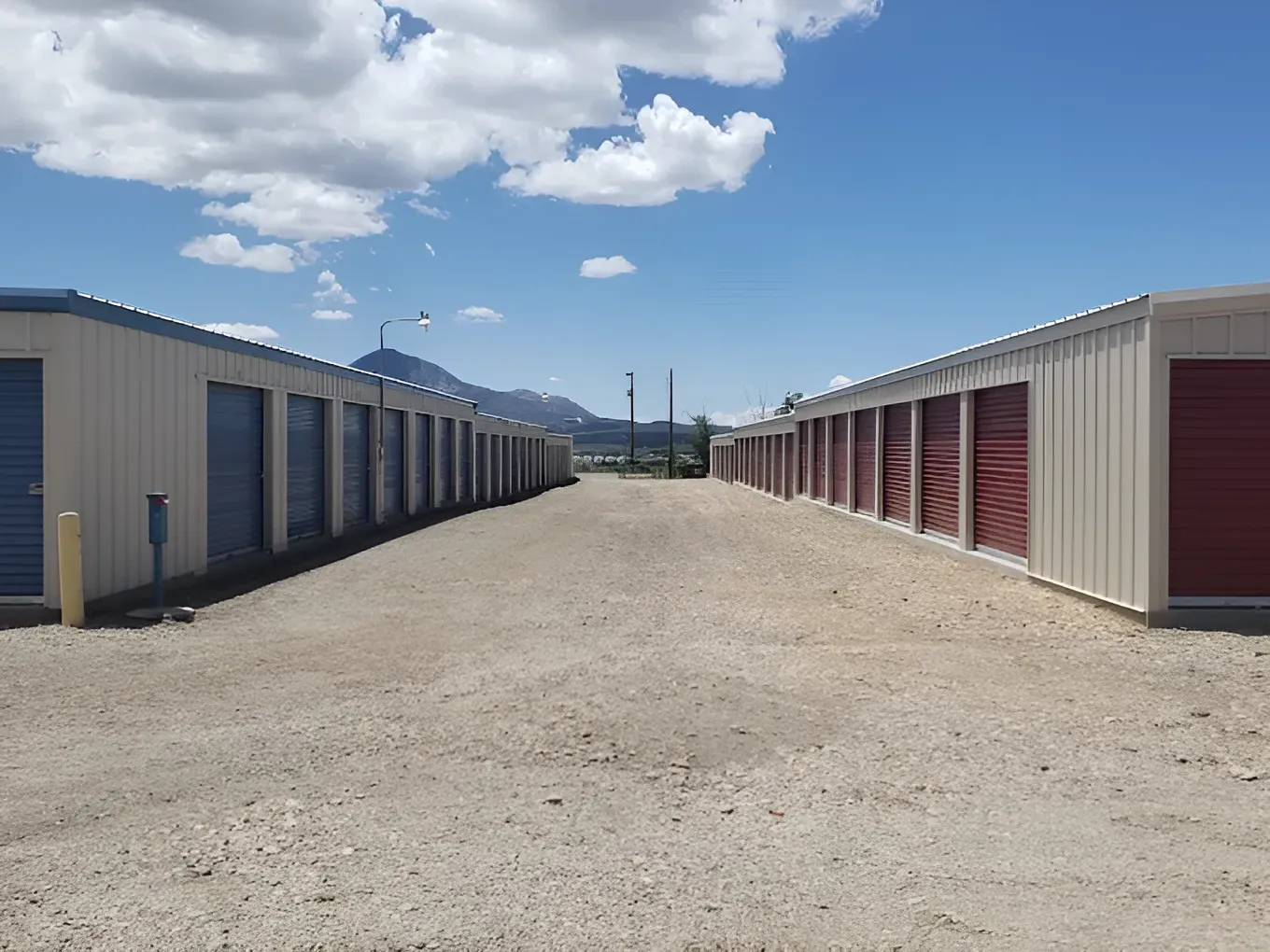 Rows of storage units with blue and red doors, gravel lot, mountain background, bright sky.