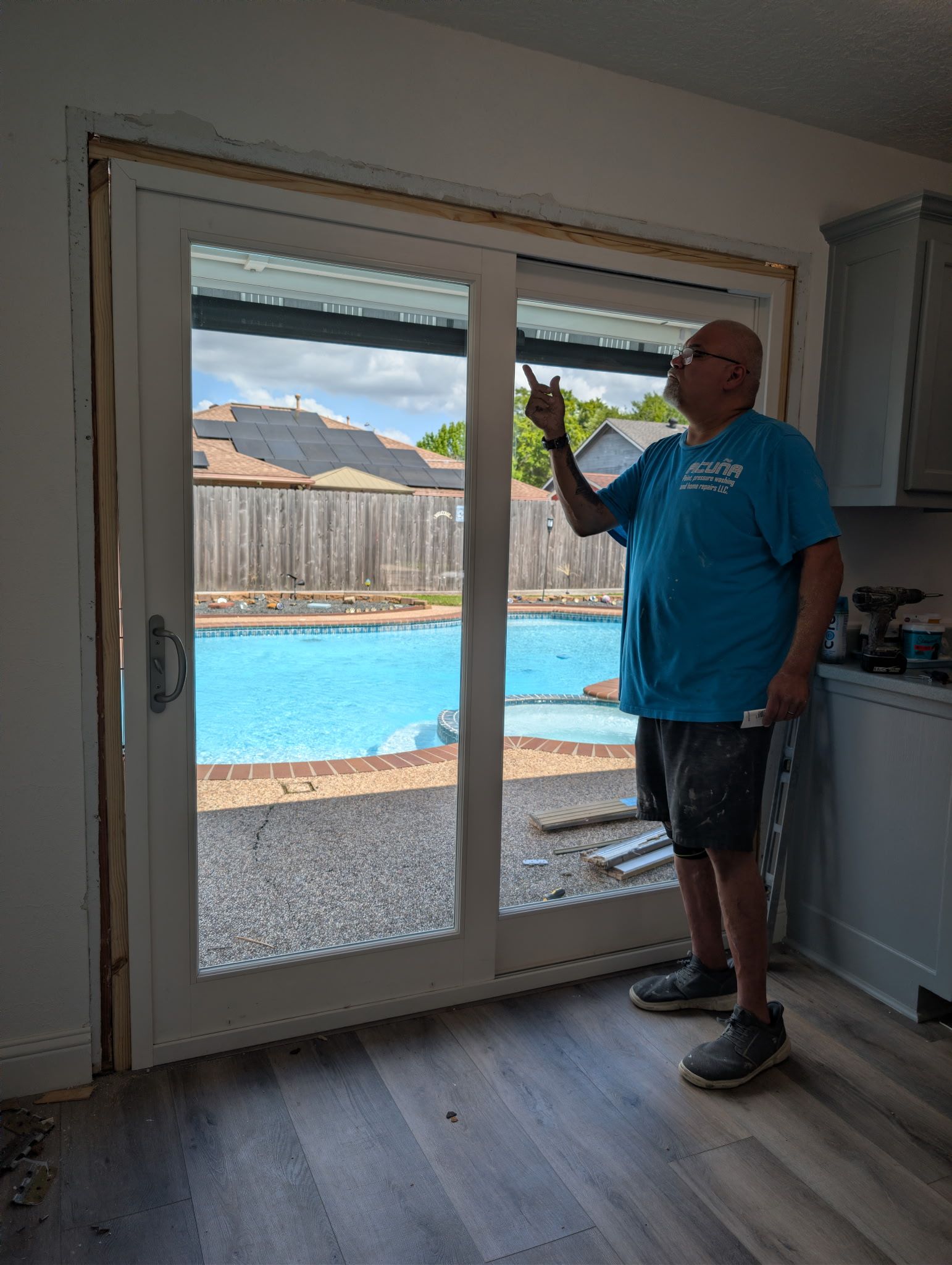 Man standing by sliding glass doors, gesturing toward a bright backyard pool and mountains beyond