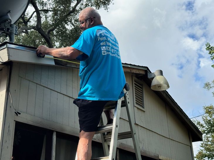 Man on roof cleaning gutters with a ladder beside a gray house under a cloudy sky