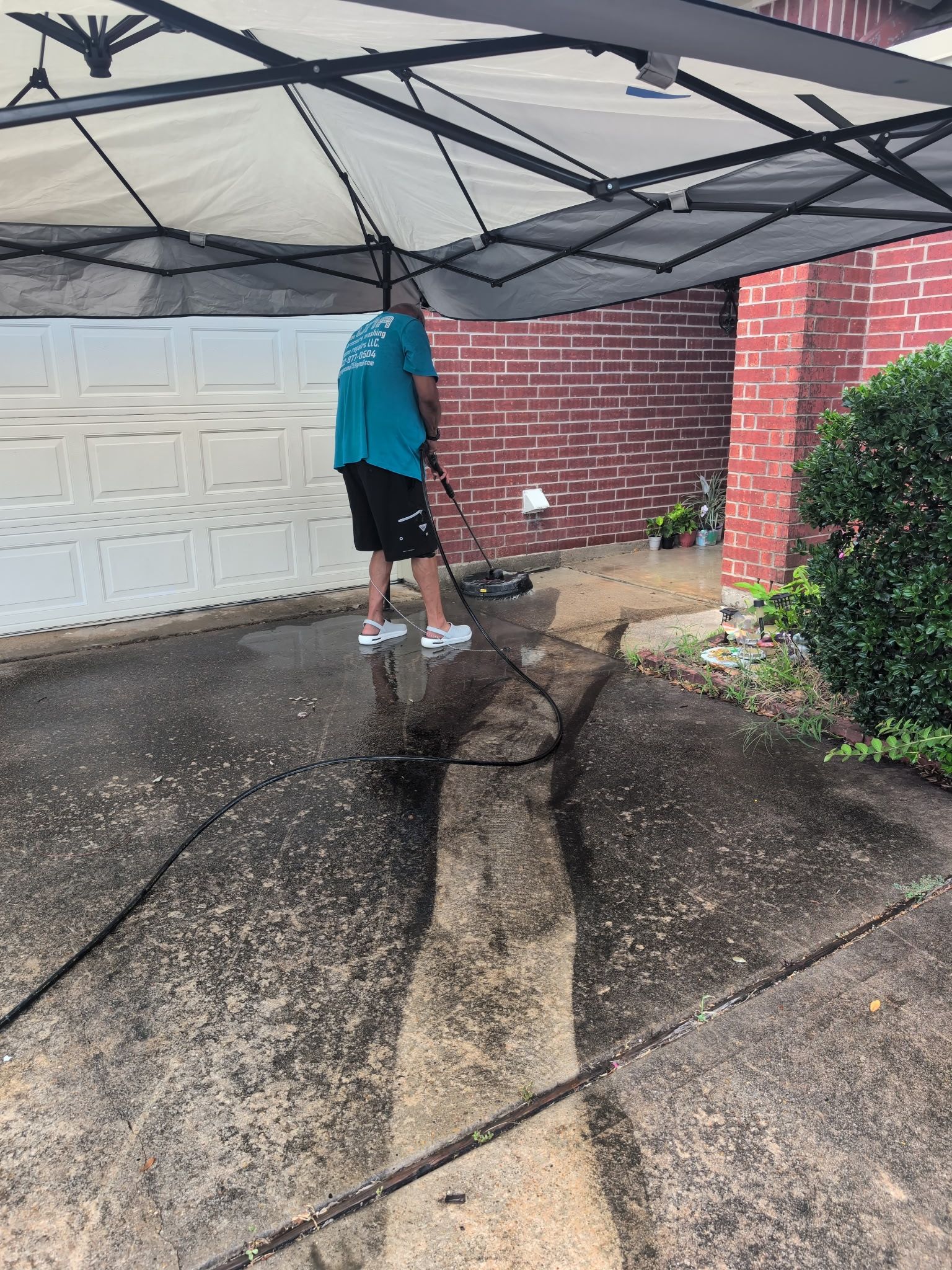 Person pressure-washing a driveway under a canopy beside a garage and brick wall