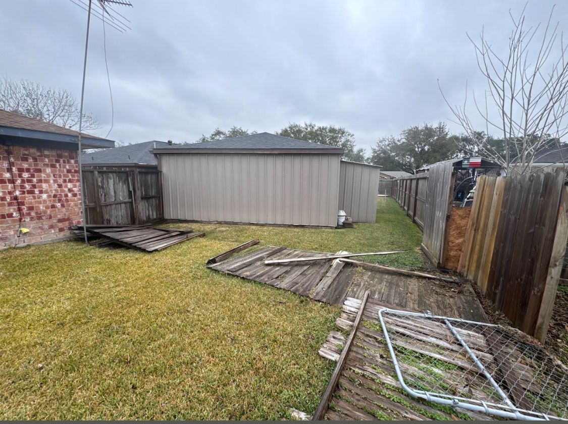 Backyard with shed, leaning fence panels, and scattered lumber on wet grass under a gray sky