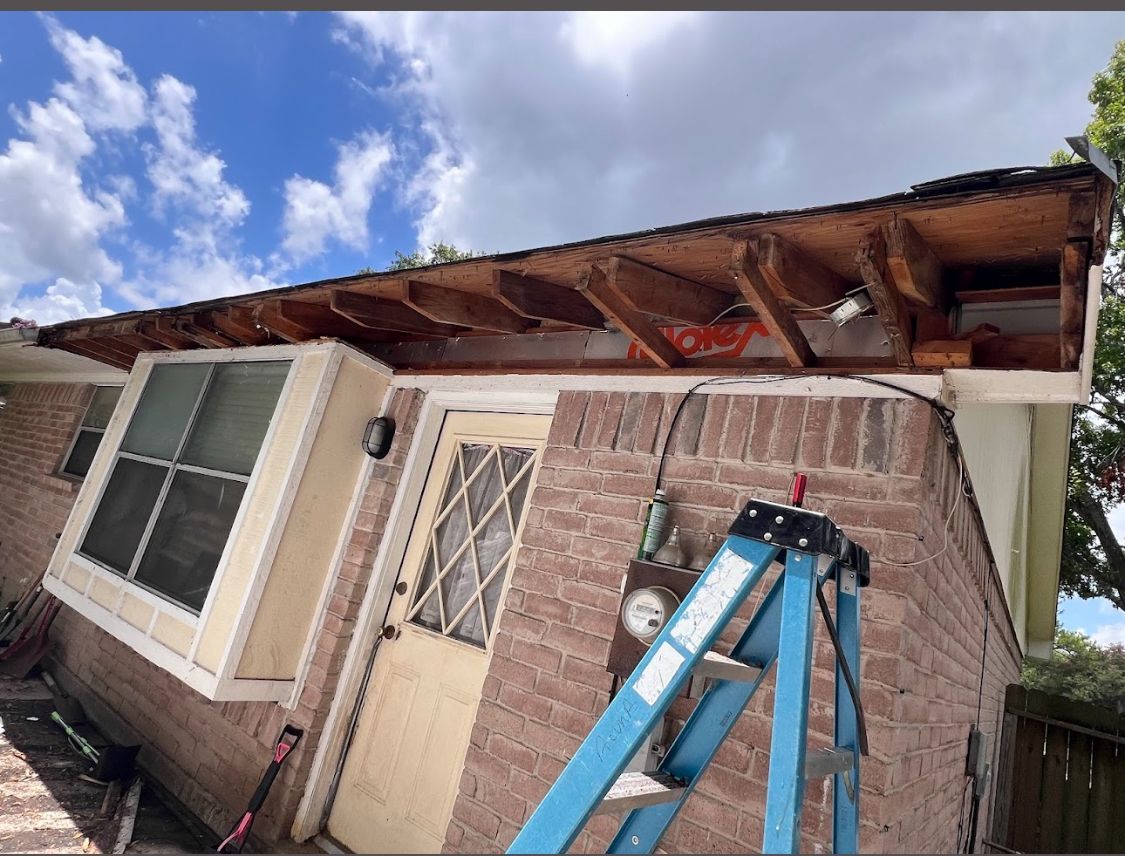 Damaged house roof with exposed beams, blue ladder, and cloudy sky above the front entrance
