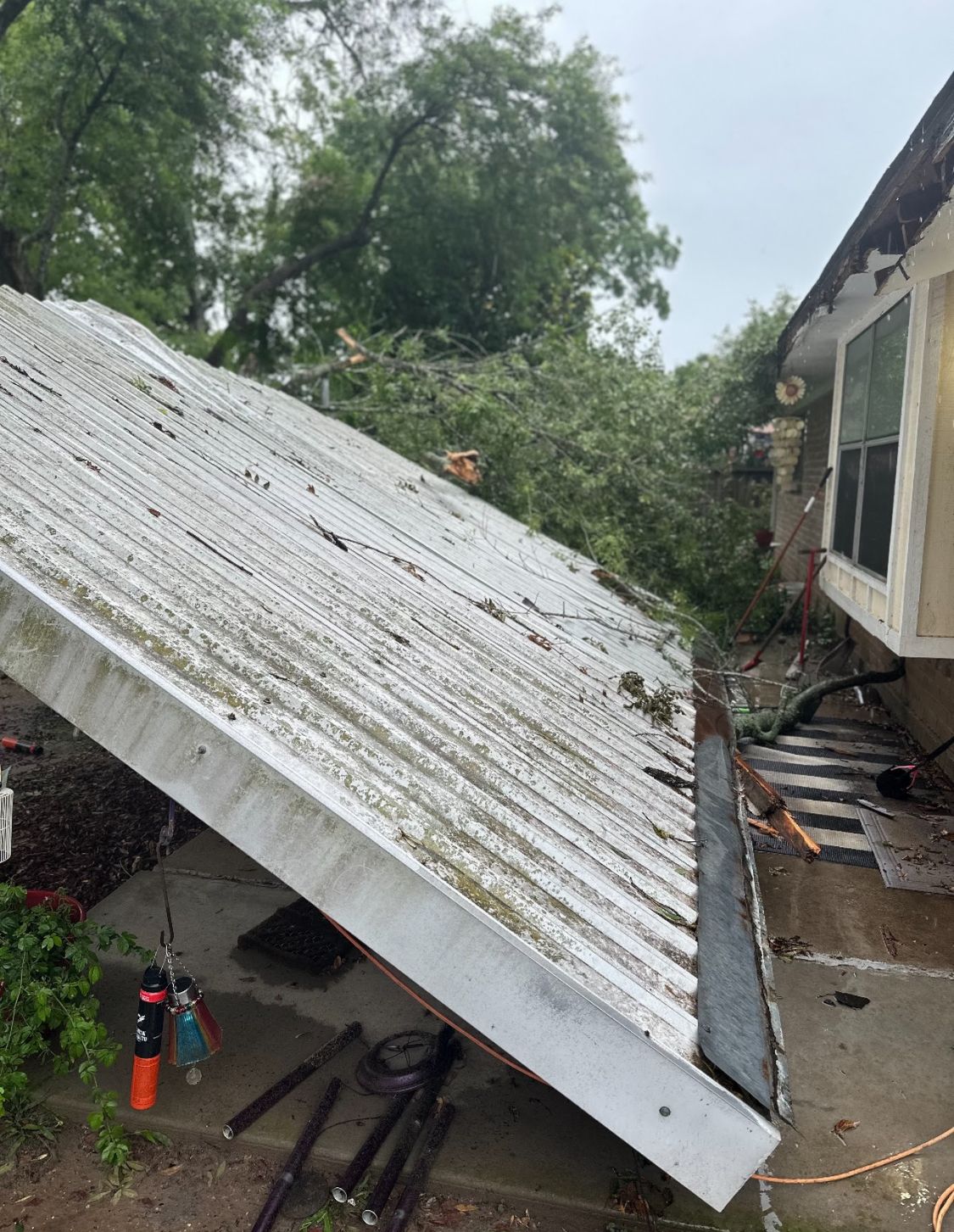 Collapsed corrugated metal roof leaning against a house in a yard, with tools and debris nearby