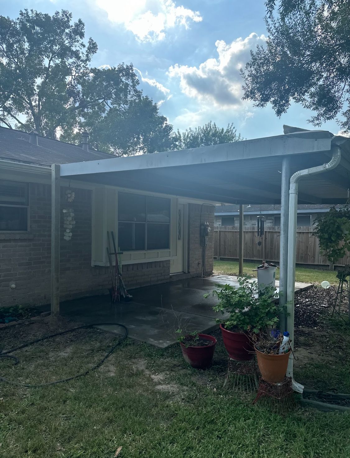 Backyard patio with a covered awning, potted plants, and brick house under a partly cloudy sky