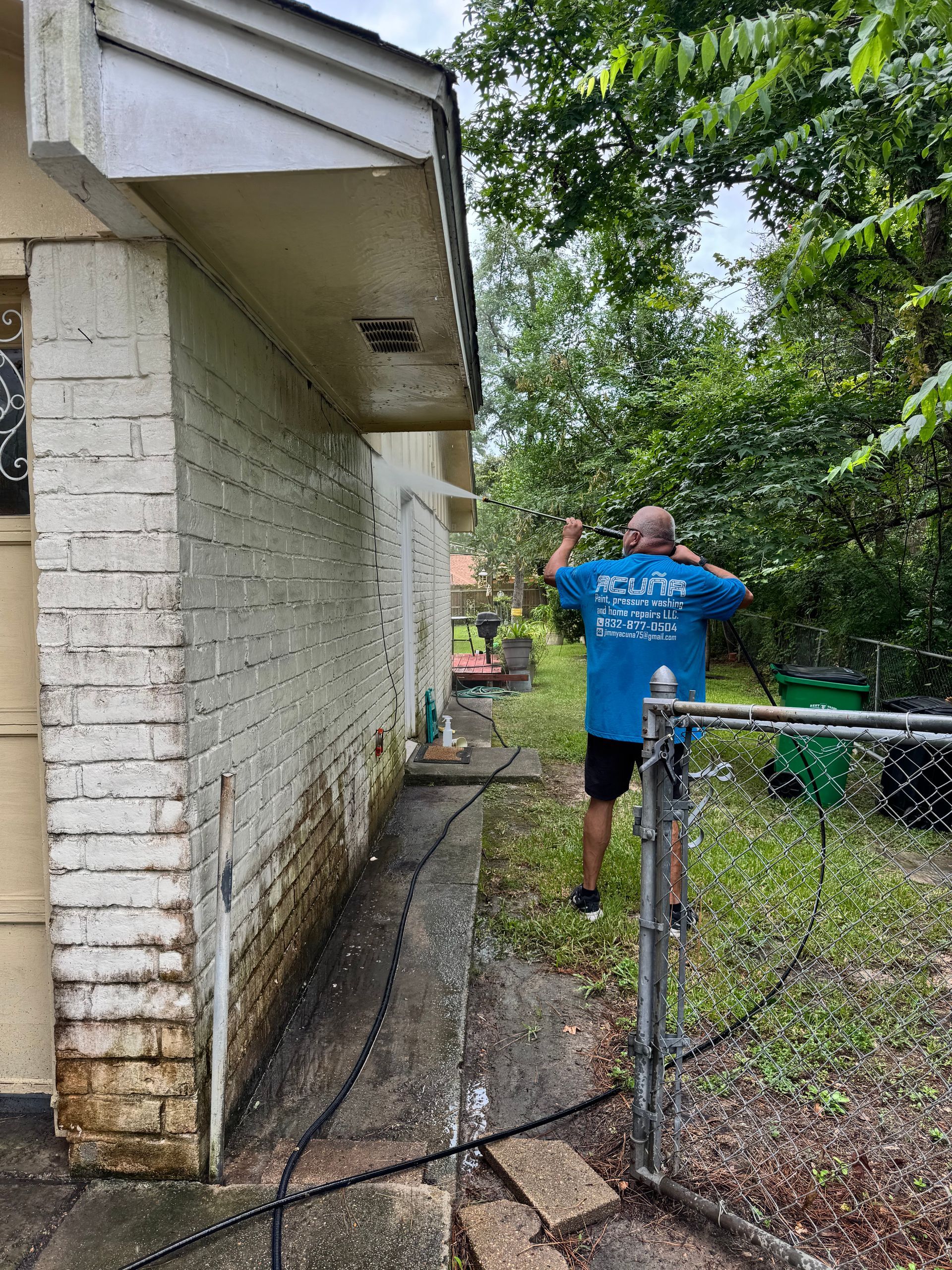 Person in blue shirt walking along a narrow path beside a brick house and chain-link fence.
