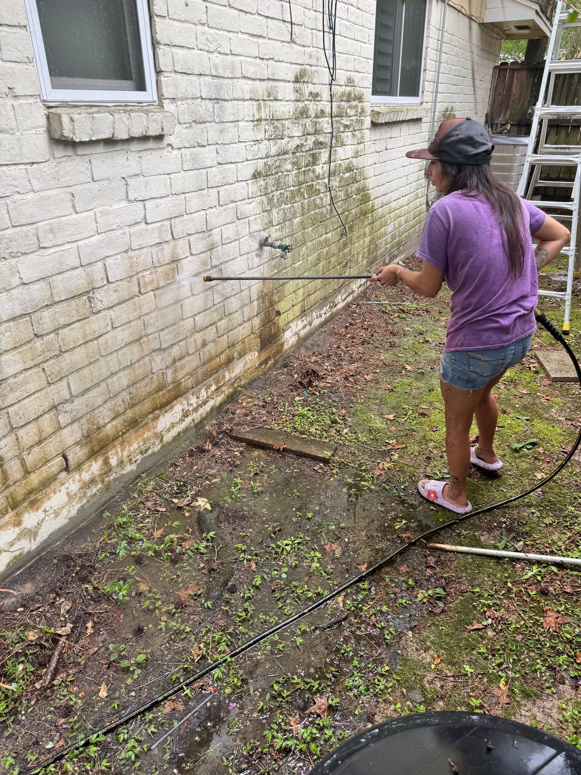 Person pressure-washing a stained white brick wall beside a muddy yard.