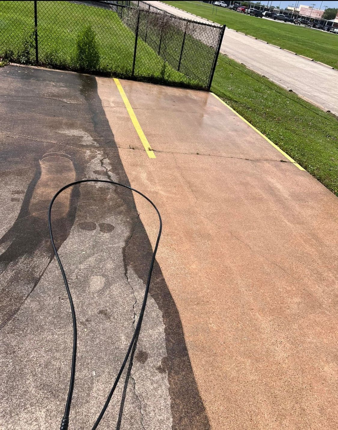 Cracked asphalt driveway beside a grassy yard, with a yellow line and chain-link fence ahead.