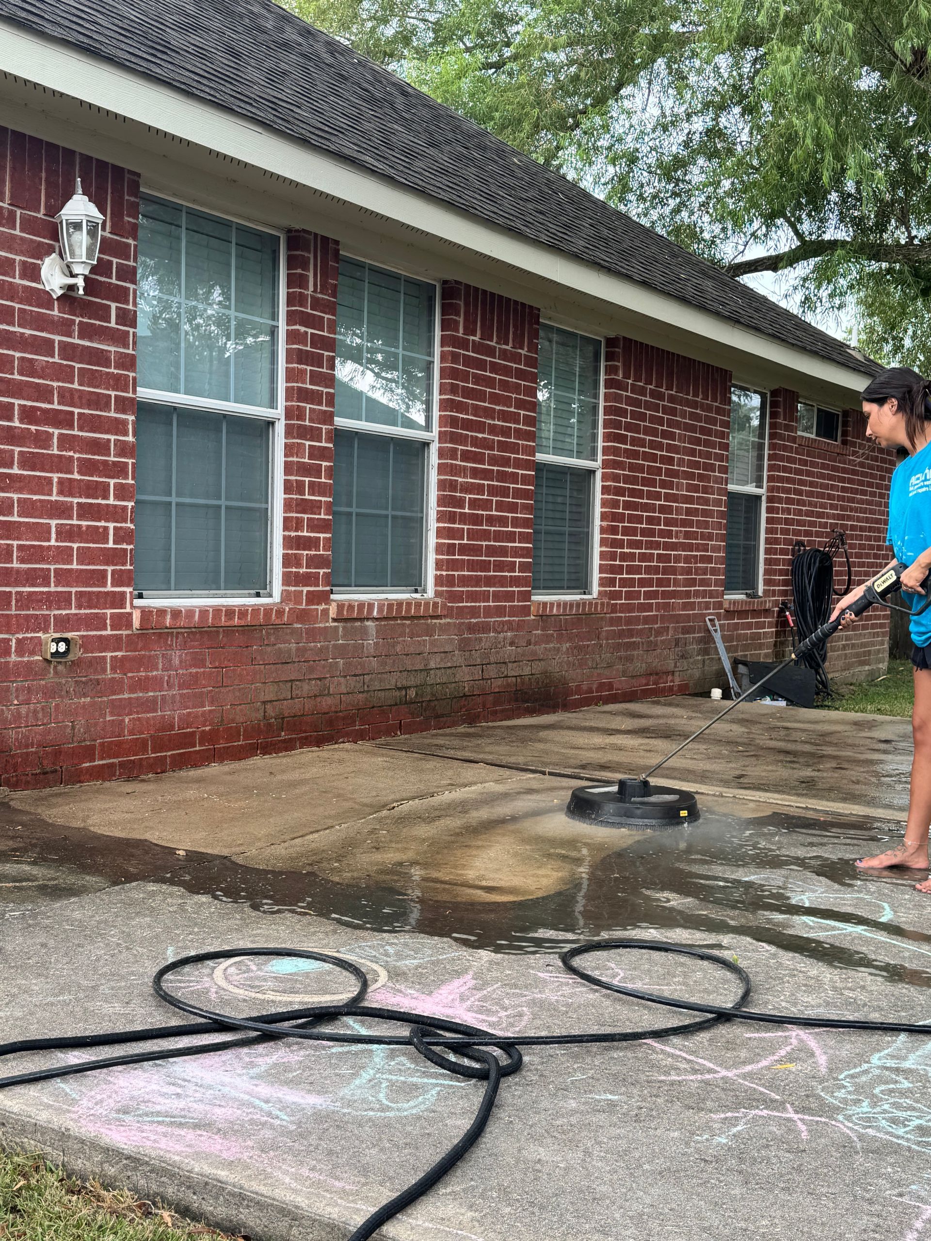 Person pressure-washes a brick house exterior near wet pavement and tangled hoses.