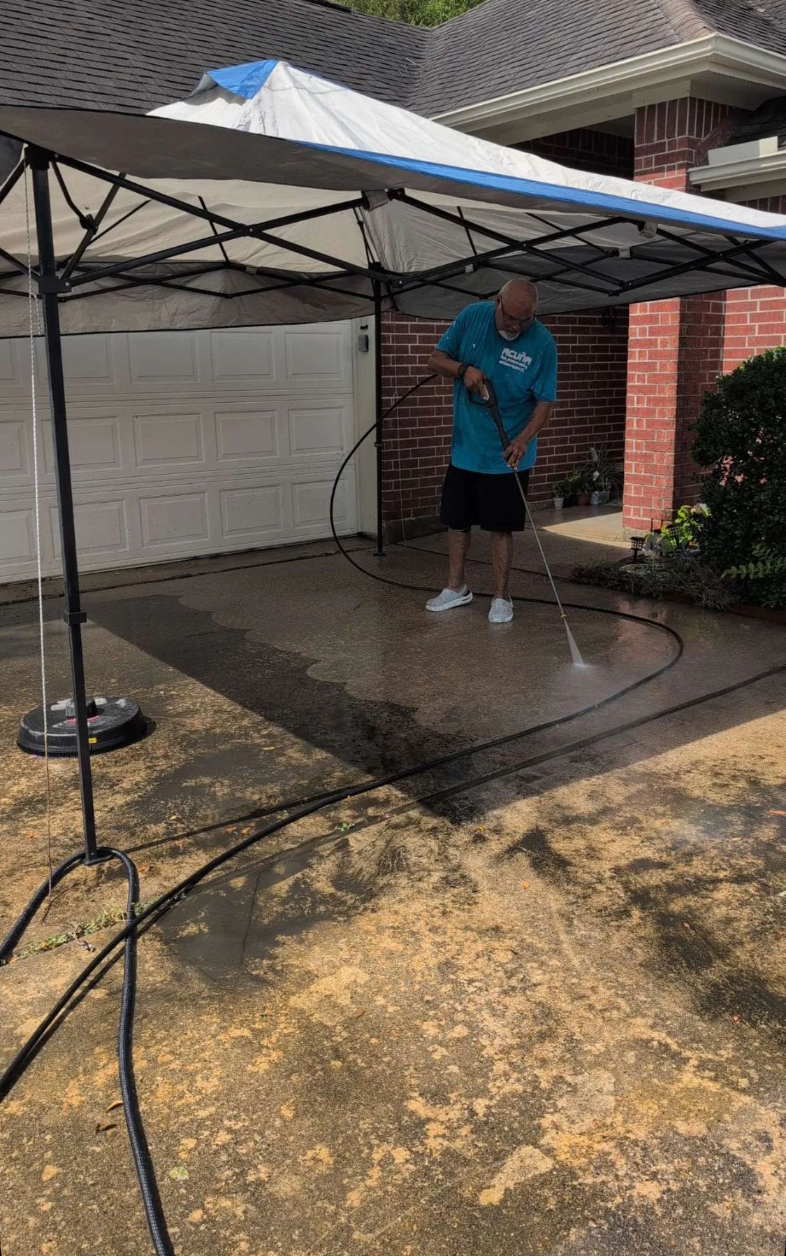 Person pressure-washing a driveway under a white canopy in front of a brick garage.
