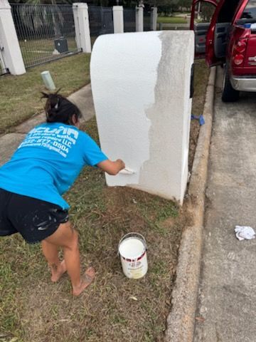 Person painting a white wall panel outdoors beside a driveway, with a paint bucket nearby.