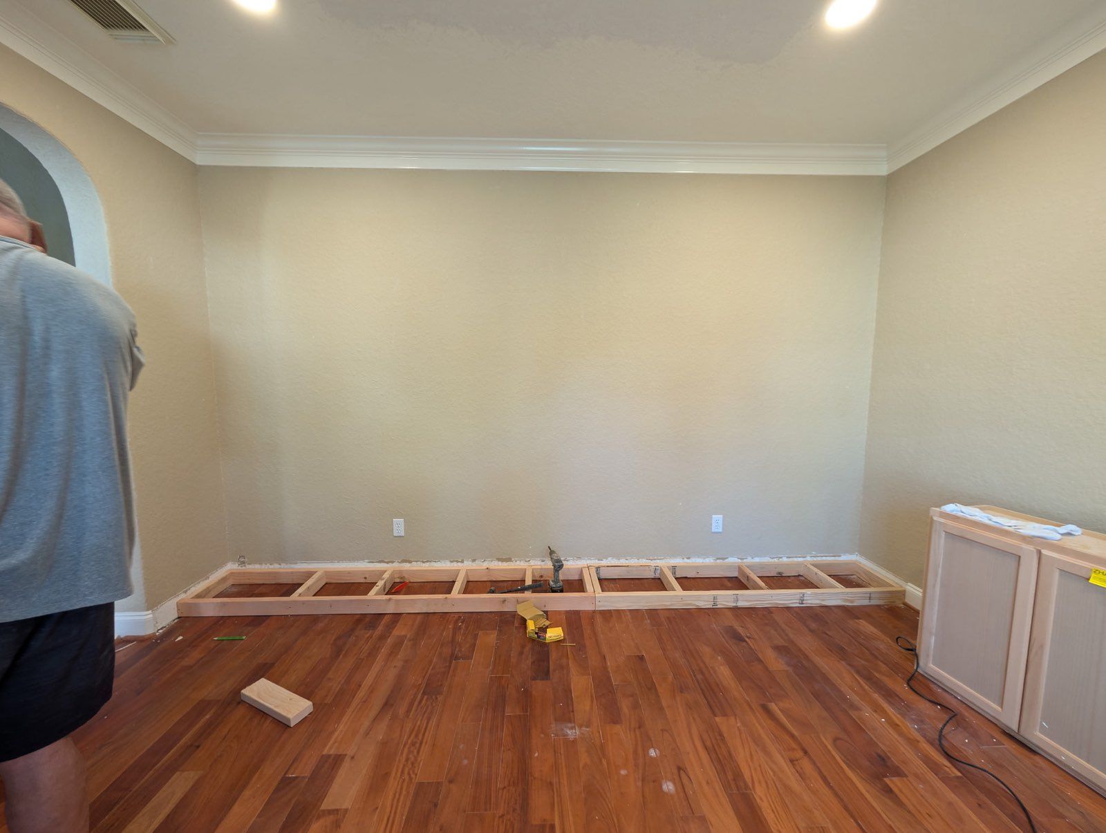 Empty room under renovation with hardwood floors, beige walls, and baseboards removed; person partially visible at left.
