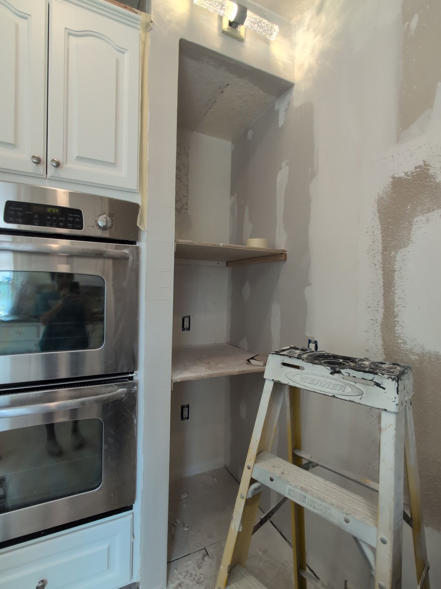 Kitchen alcove under renovation with patchy drywall, shelves, and a step ladder beside built-in ovens