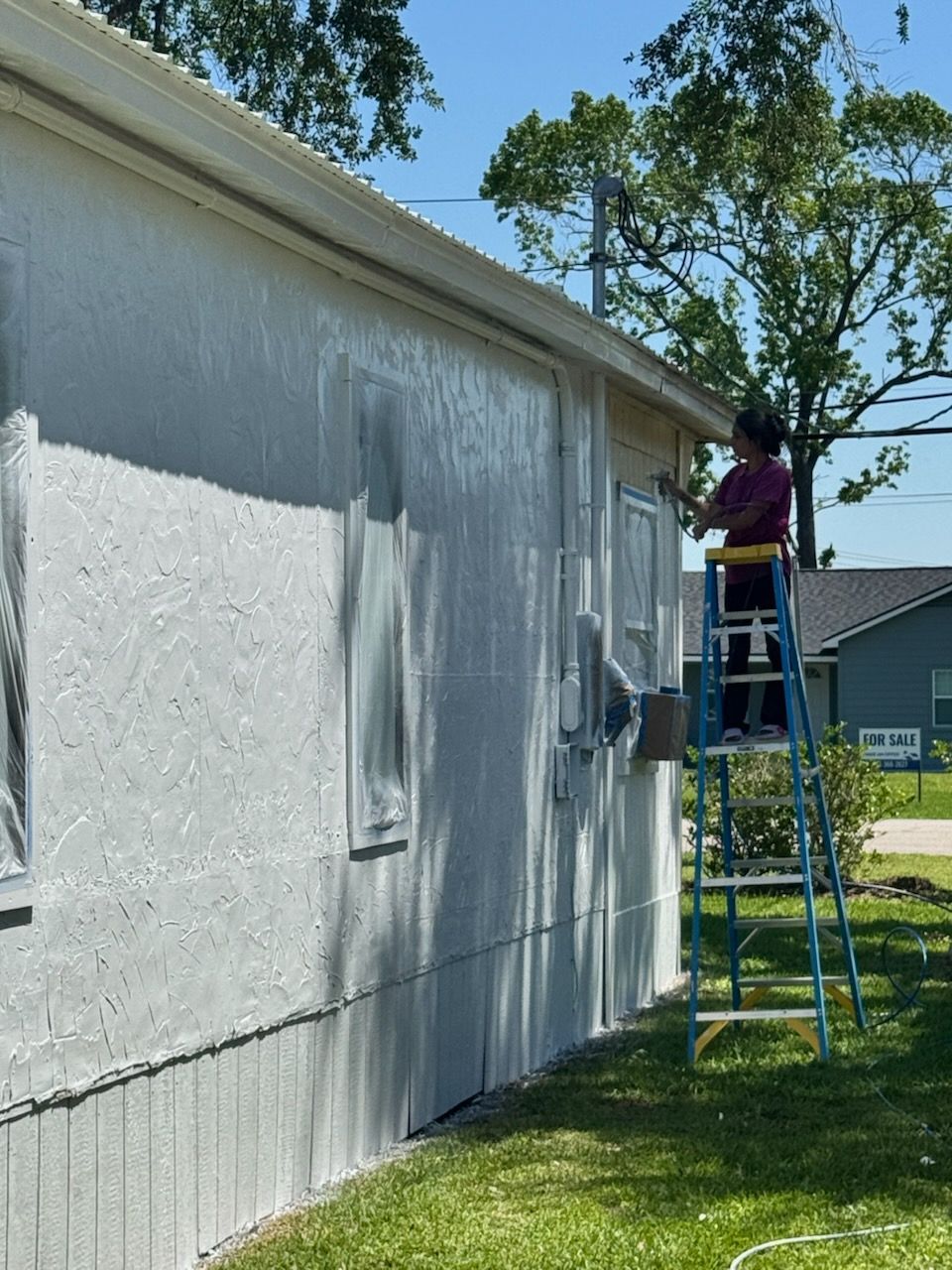 Person painting the side of a white house while standing on a ladder in the yard