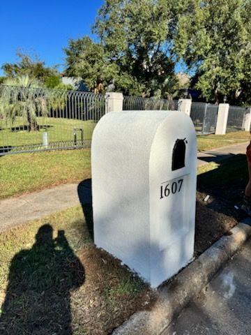 White mailbox numbered 1607 beside a curb, with a grassy yard and fence in sunlight.