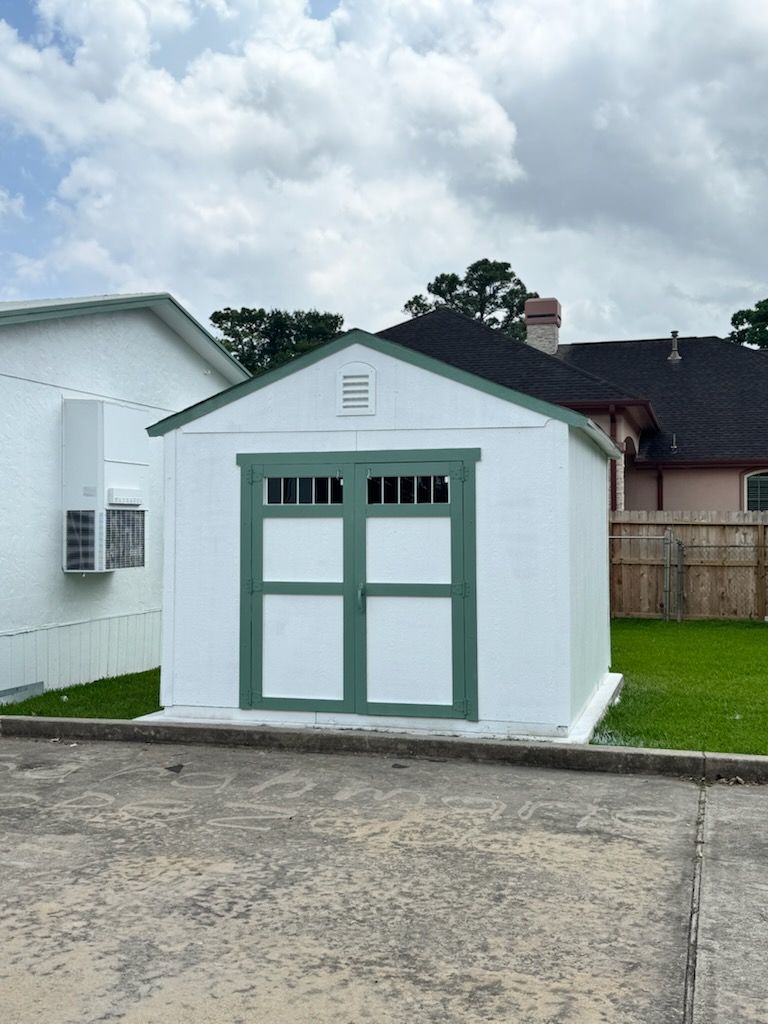 Small white shed with green-trimmed double doors in a driveway, under a cloudy sky.
