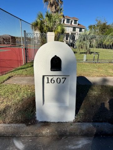 White mailbox with house number 1607 beside a tennis court and grass, in front of a house.