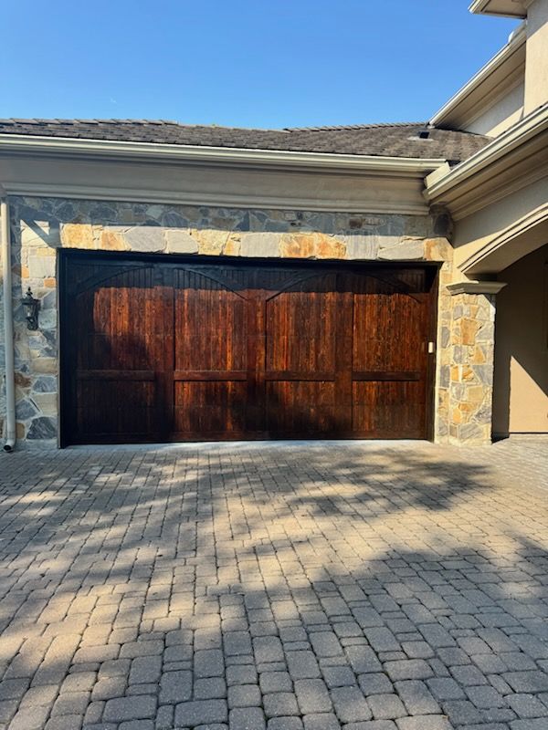 Stone-framed dark wood garage door beneath a beige roof, with a paved driveway in front