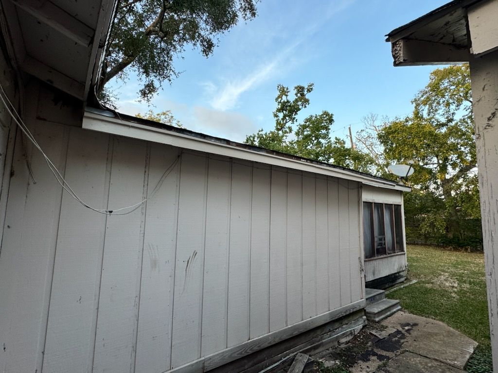 Exterior of a white house with cracked siding and a side window, viewed along a narrow yard