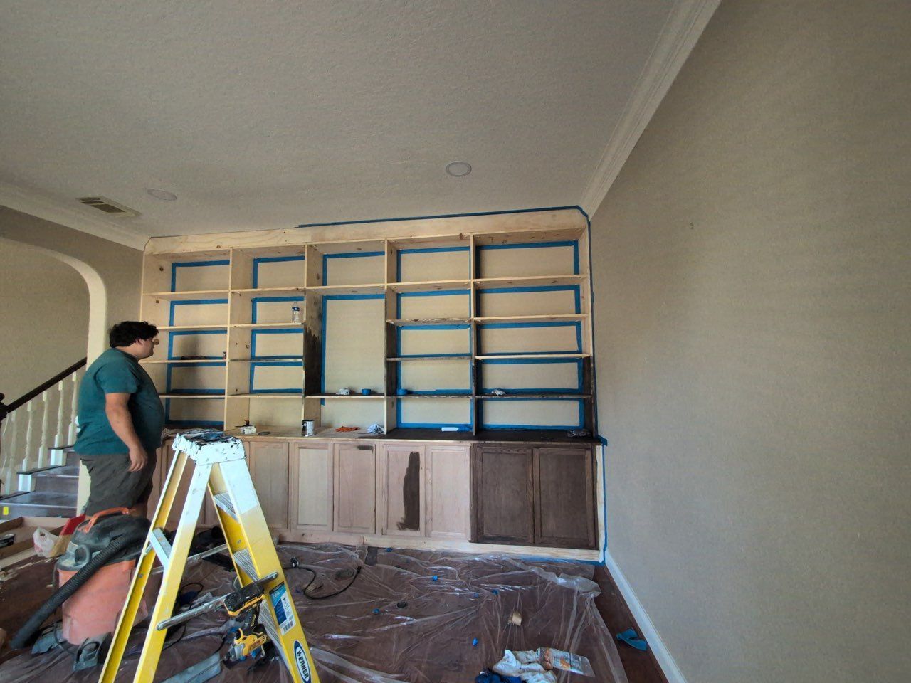 Worker painting built-in shelves and cabinets in a room, with a yellow ladder in the foreground.