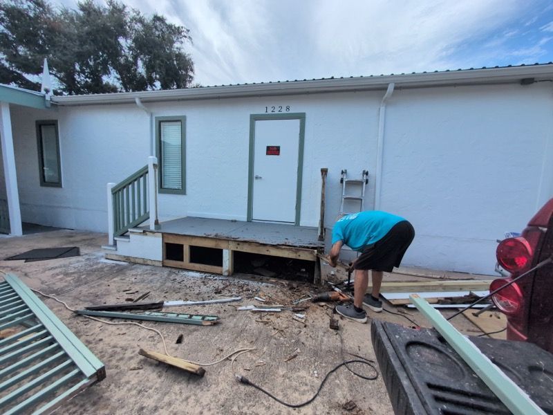 Person working on a damaged porch outside a white house, with debris and tools on the ground.