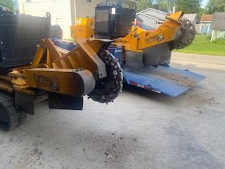A yellow stump grinder is parked next to a blue trailer.