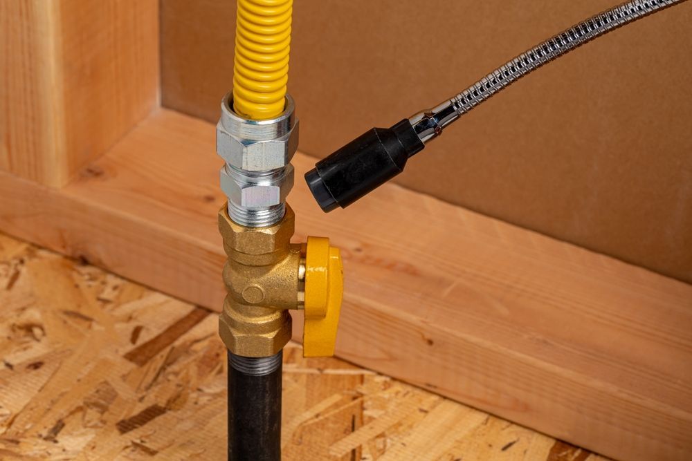 A close-up of a yellow gas shut-off valve on a pipe, with a flexible metal detector nozzle pointed toward the connection.