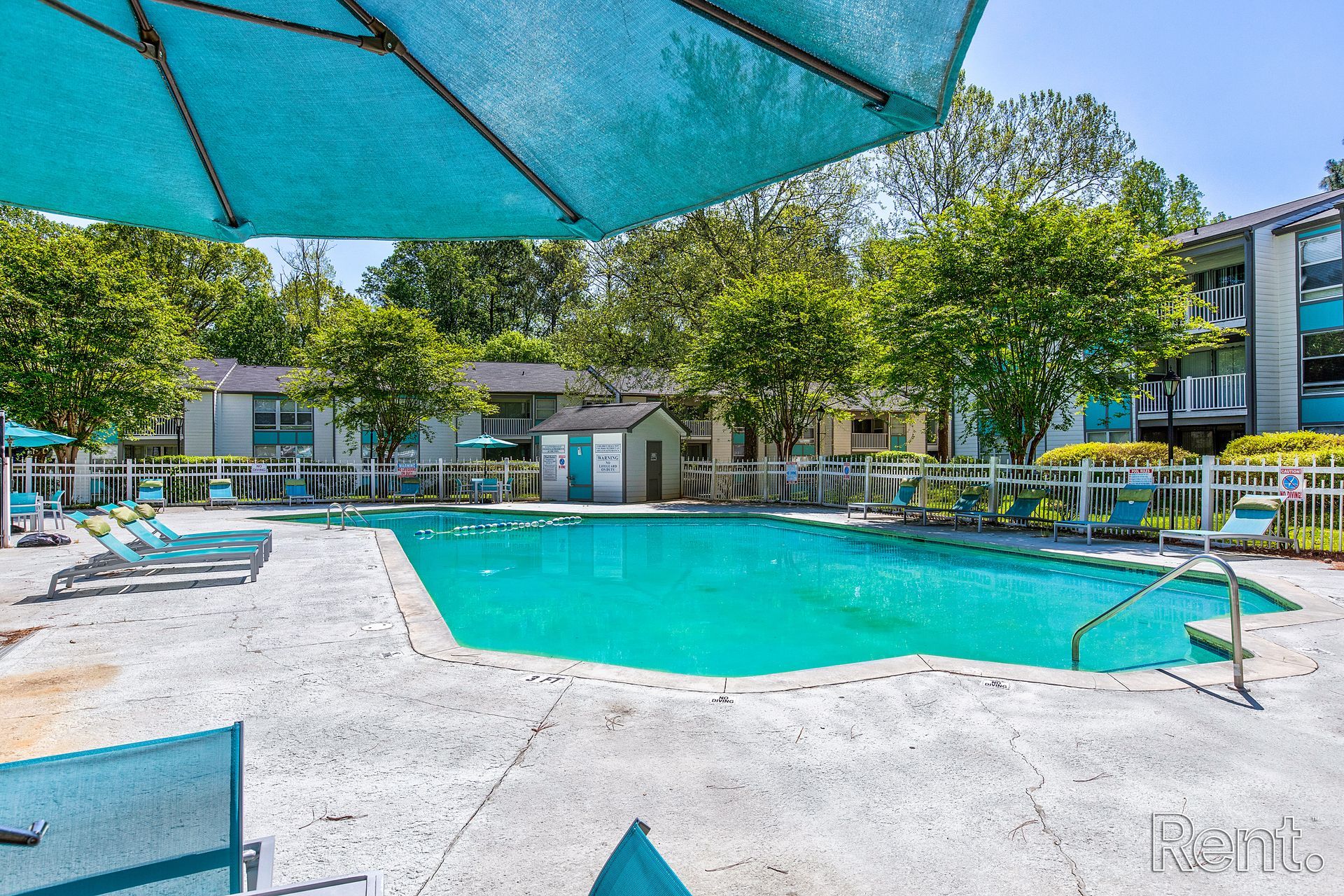 Outdoor apartment pool with turquoise water, lounge chairs, trees, and blue umbrellas under a sunny sky