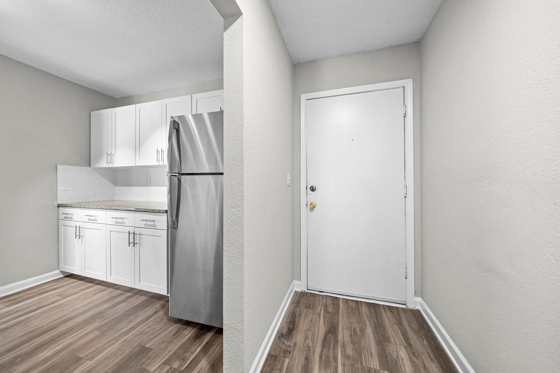 Small apartment entryway beside a kitchen with white cabinets, stainless fridge, and wood floors.