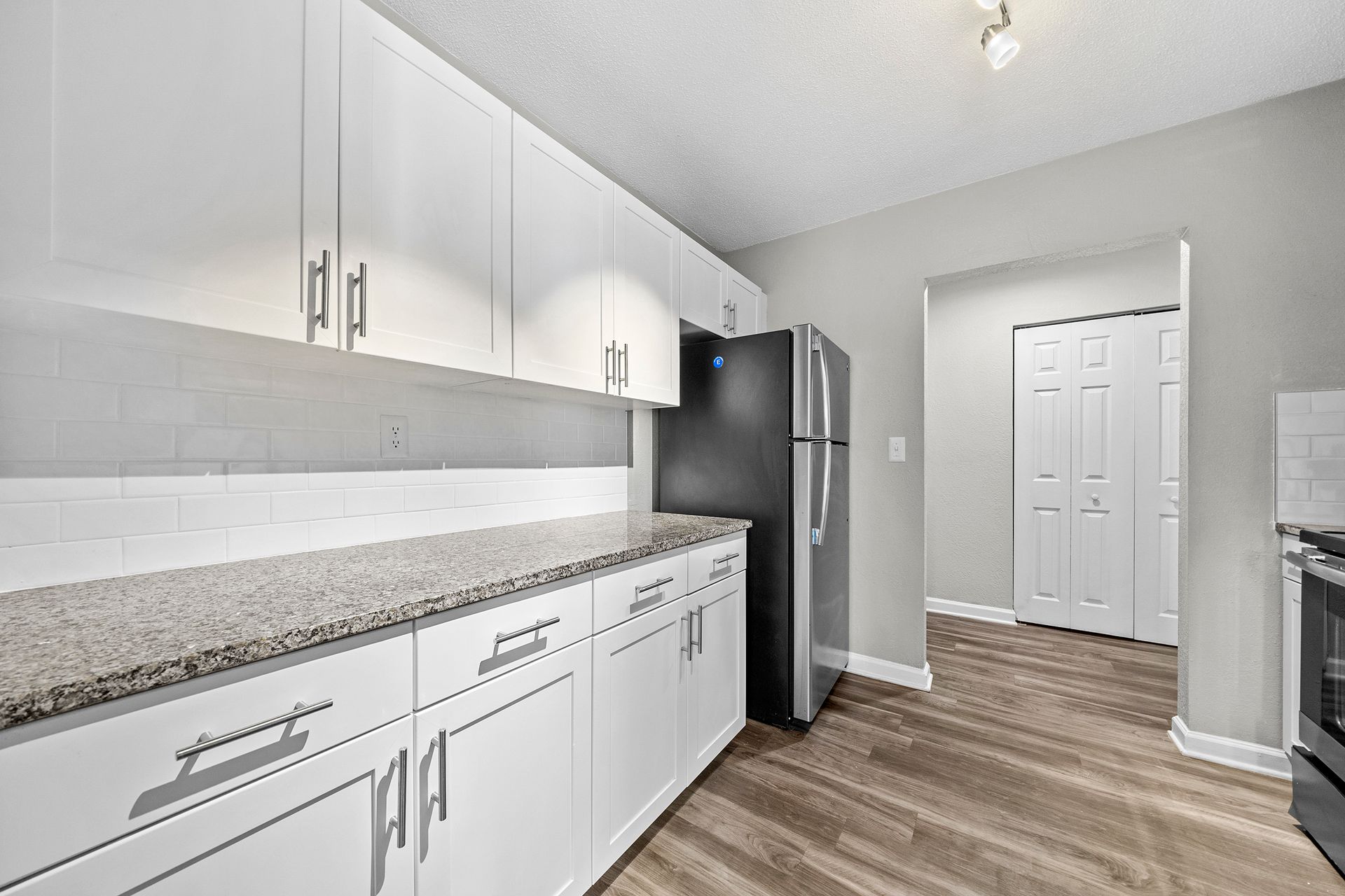 Bright modern kitchen with white cabinets, granite counters, stainless fridge, and wood-look flooring.