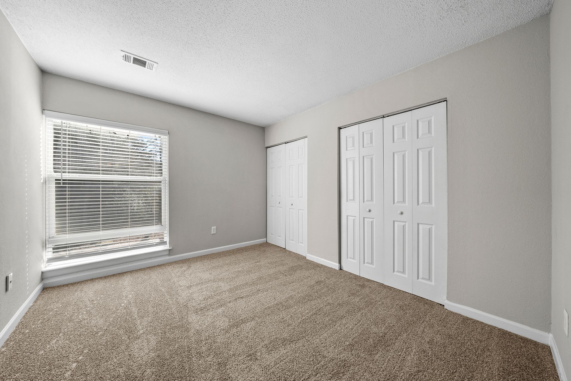 Empty beige bedroom with carpet, white closets, and a window with blinds
