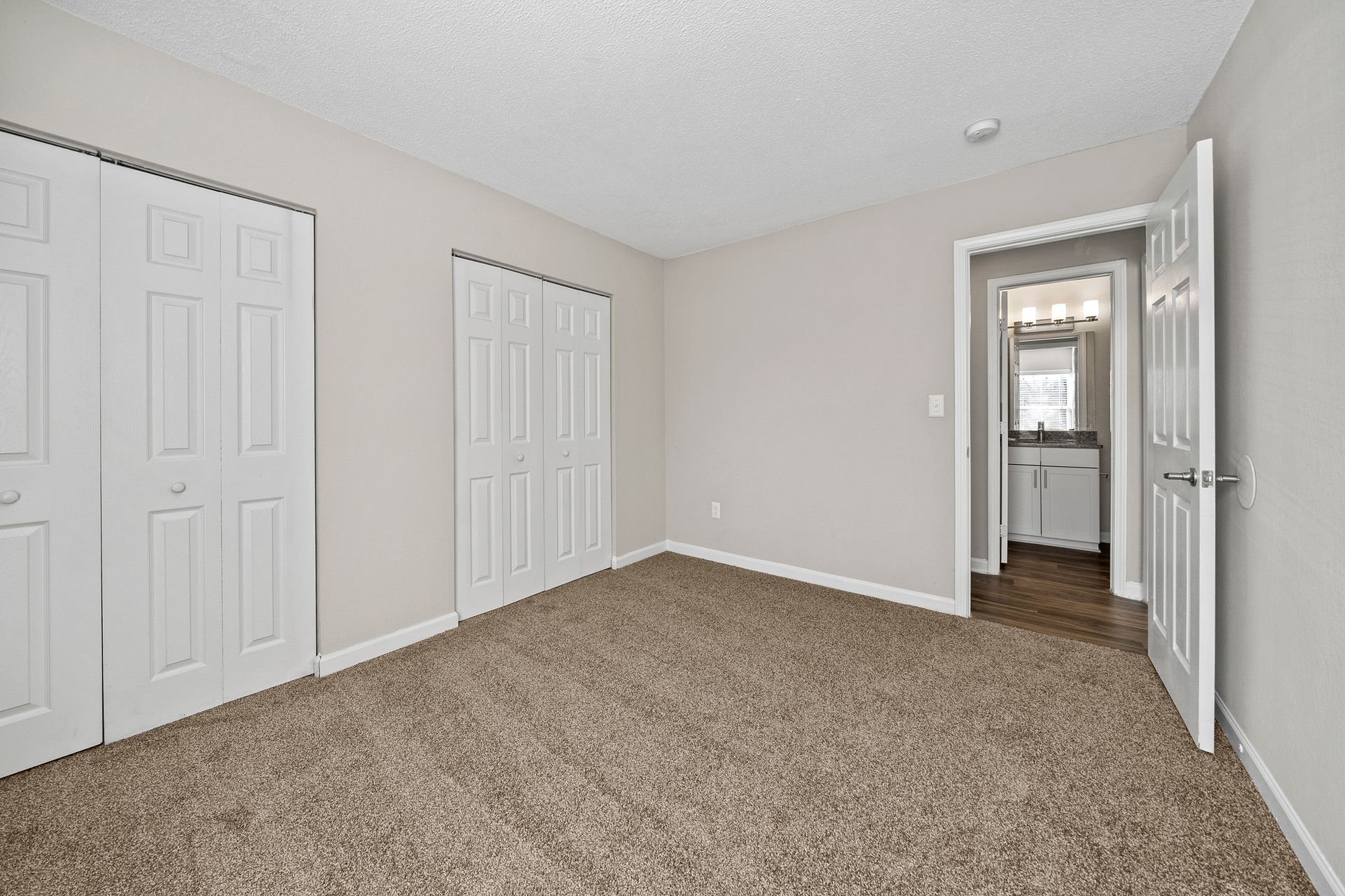 Empty carpeted bedroom with beige walls, white closet doors, and an open door to a bathroom.