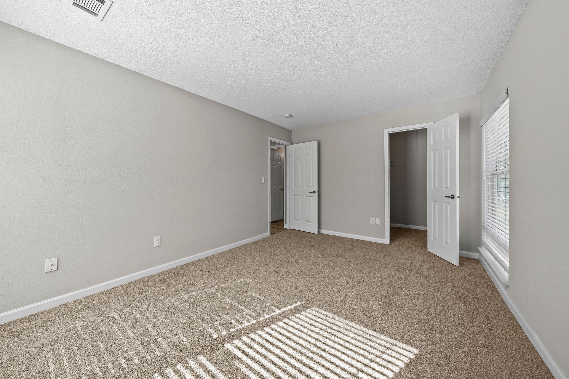 Empty beige bedroom with carpet, gray walls, open doors, and sunlight through blinds.