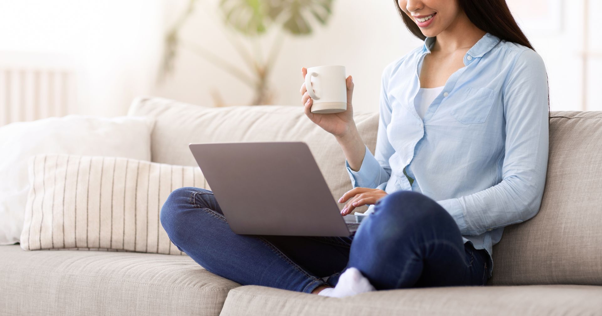 Person sitting on a couch with a laptop and holding a mug in a bright living room