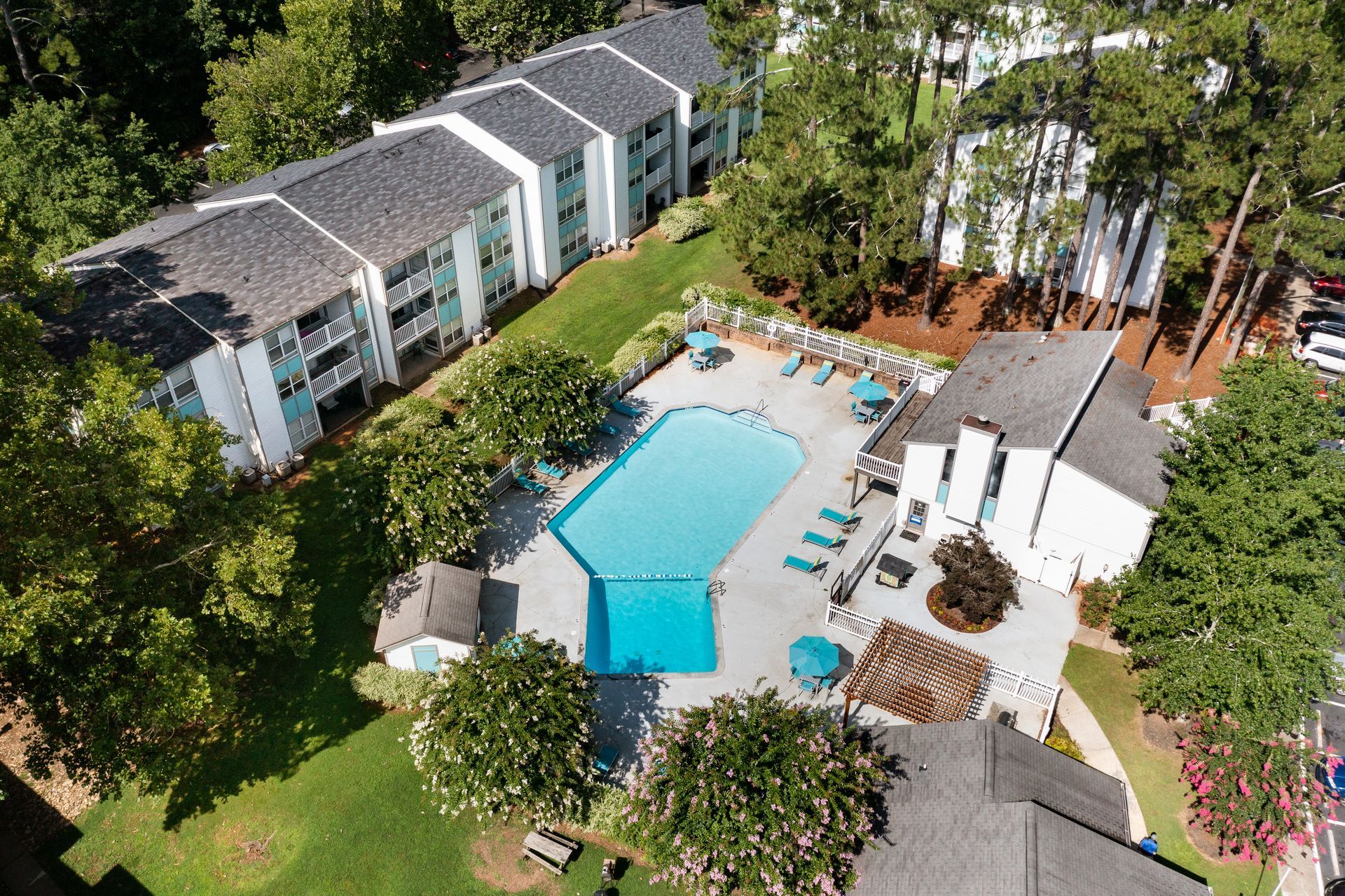 Aerial view of a resort pool surrounded by lounge chairs, umbrellas, and trees beside apartment buildings