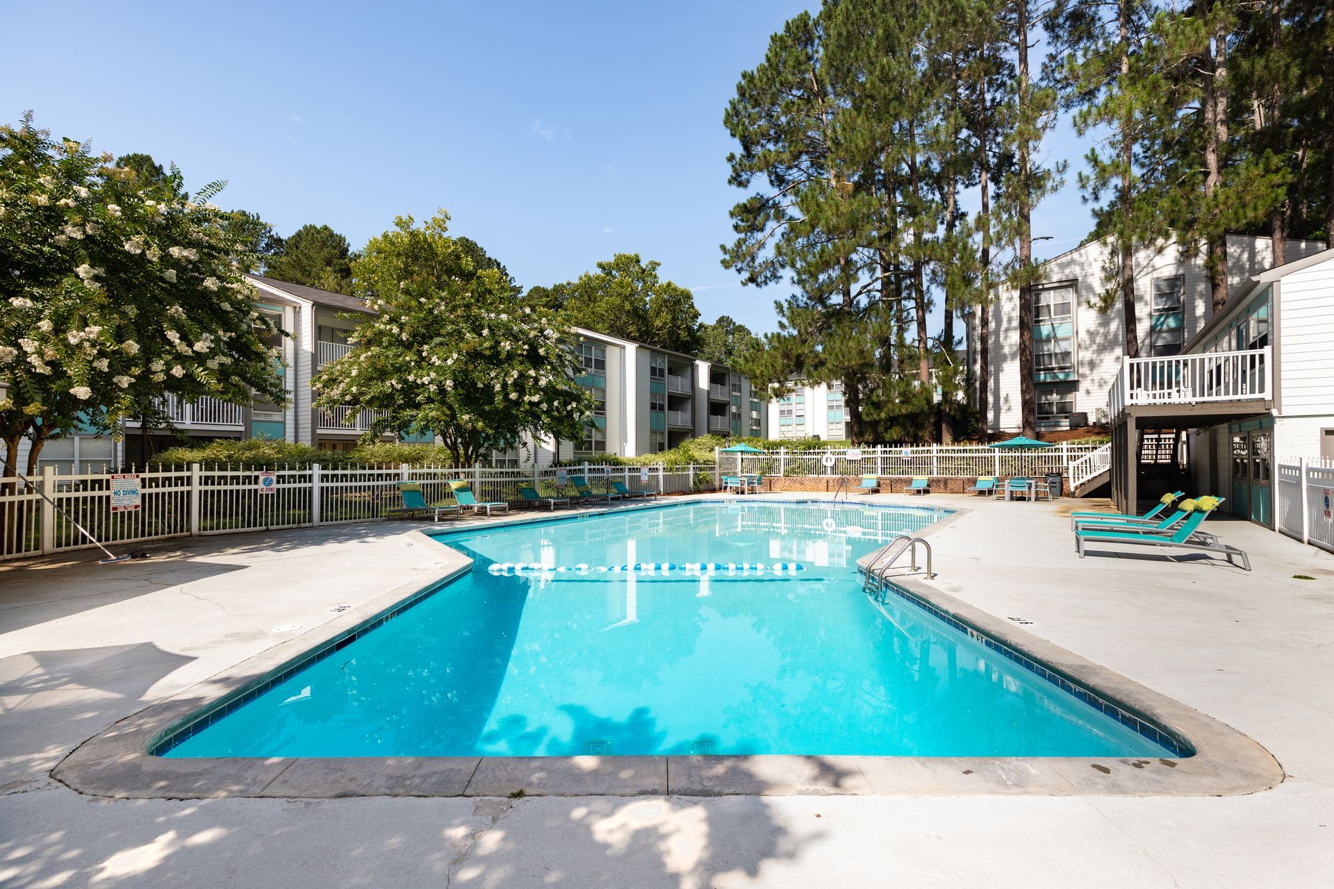 Outdoor apartment complex swimming pool with blue water, lounge chairs, and trees on a sunny day