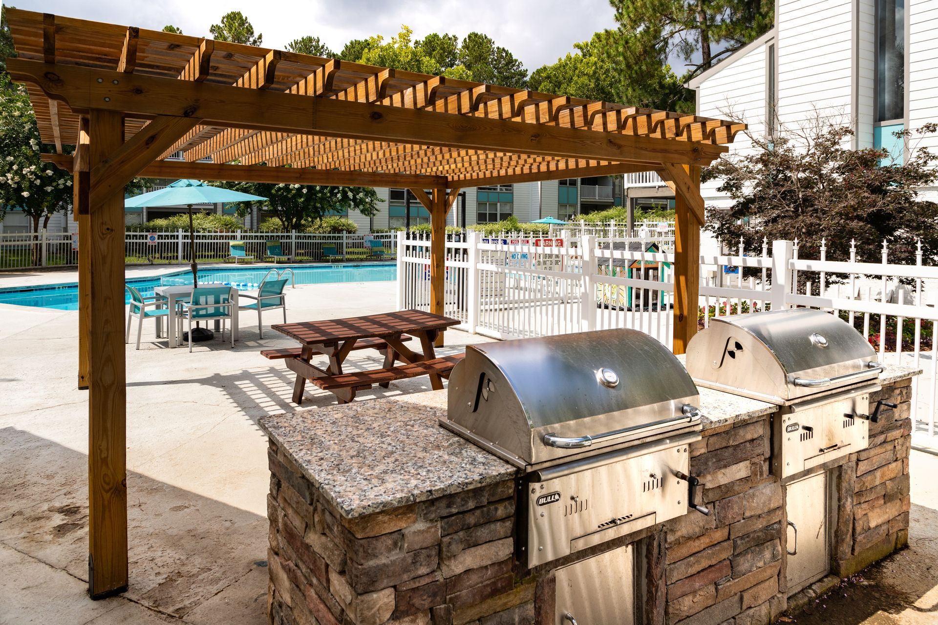 Poolside barbecue area with twin stainless grills, picnic tables, and a wooden pergola beside a swimming pool