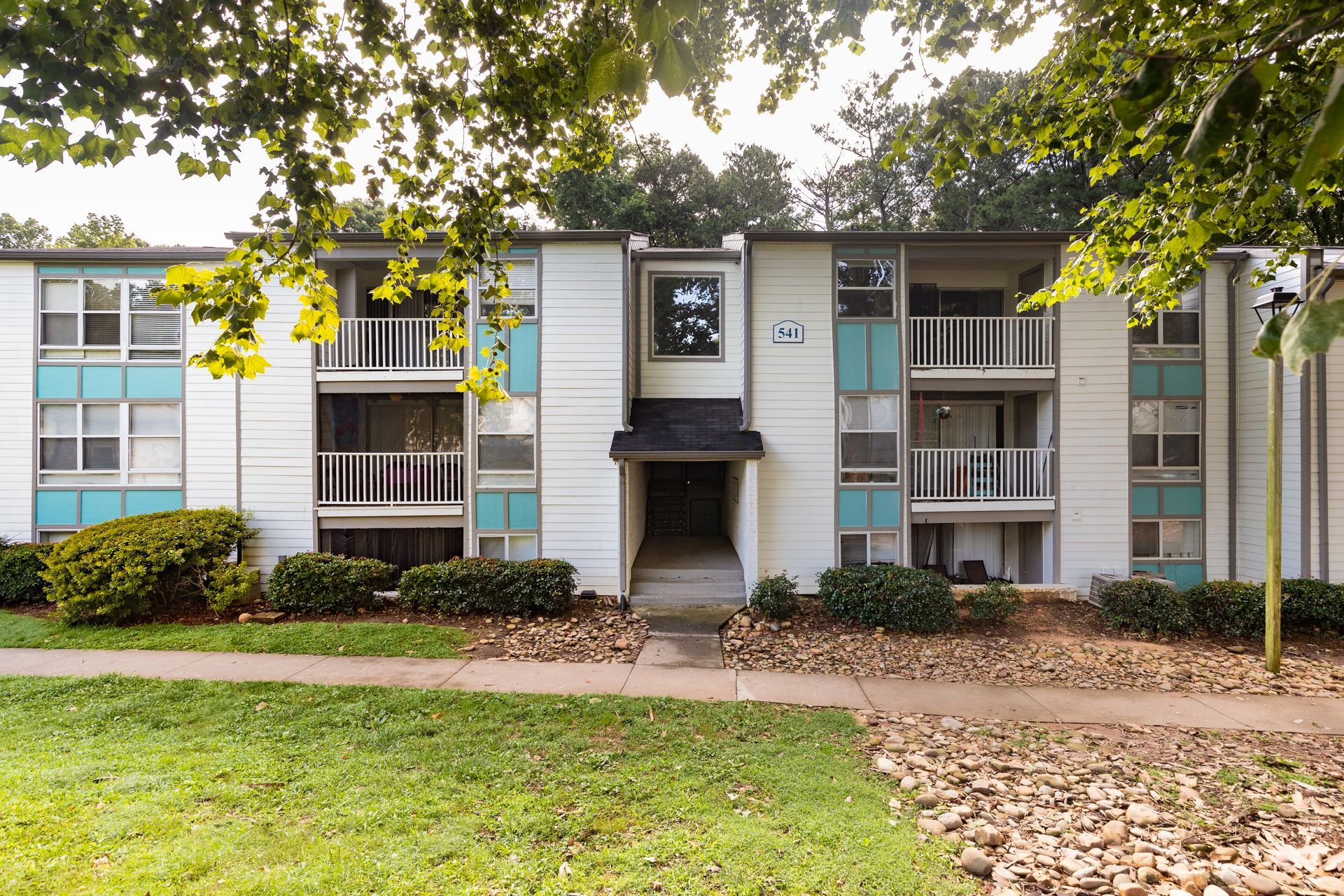 Apartment building exterior with balconies, gray siding, and a central entry walkway amid trees and lawn