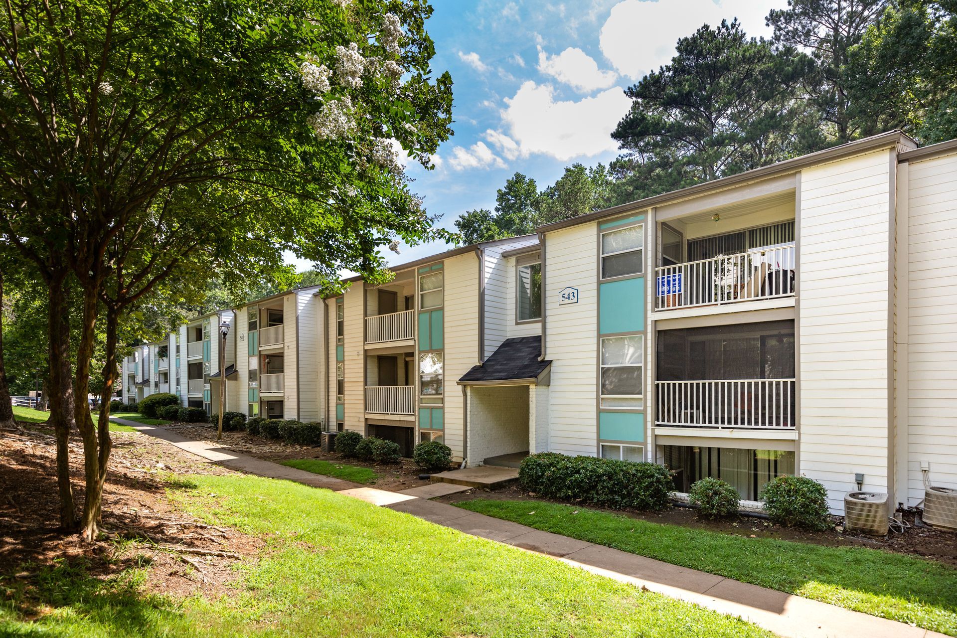 Apartment complex with balconies, bordered by trees and a grassy walkway under a blue sky