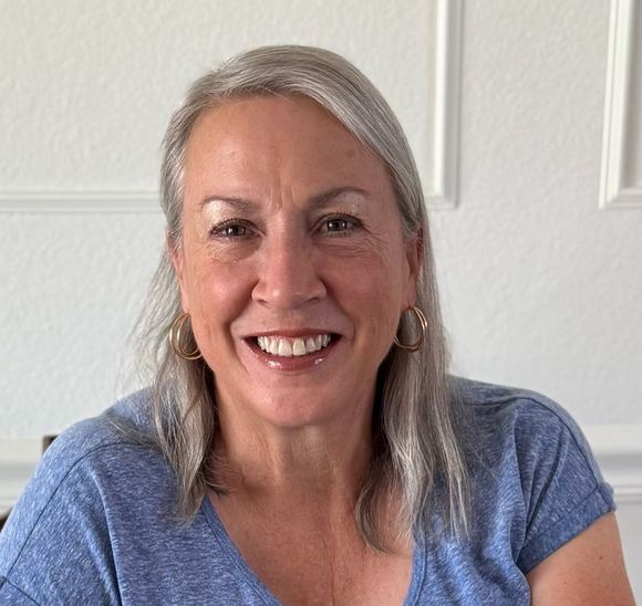 Smiling person with gray hair in a blue shirt against a white wall