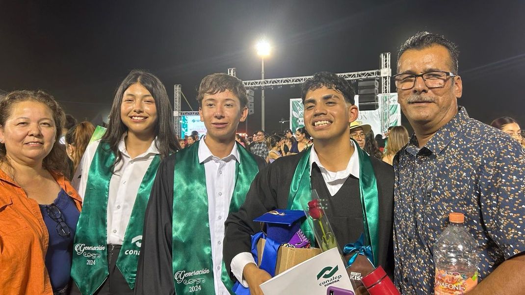 Group posing outdoors at night, with graduates in green sashes holding bouquets beside two adults.