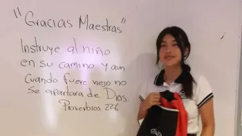 Young person standing beside a whiteboard with a handwritten “Gracias Maestros” message
