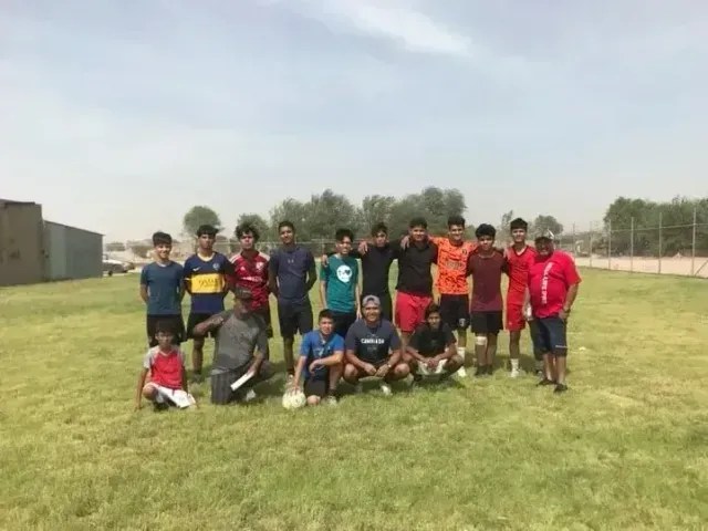Group of soccer players posing on a grassy field, with trees and a building in the background.