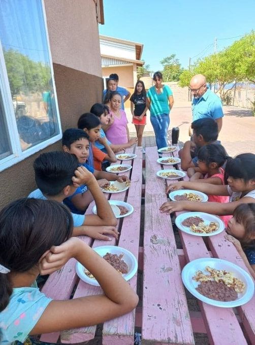 Children and adults eating lunch at long outdoor tables beside a building